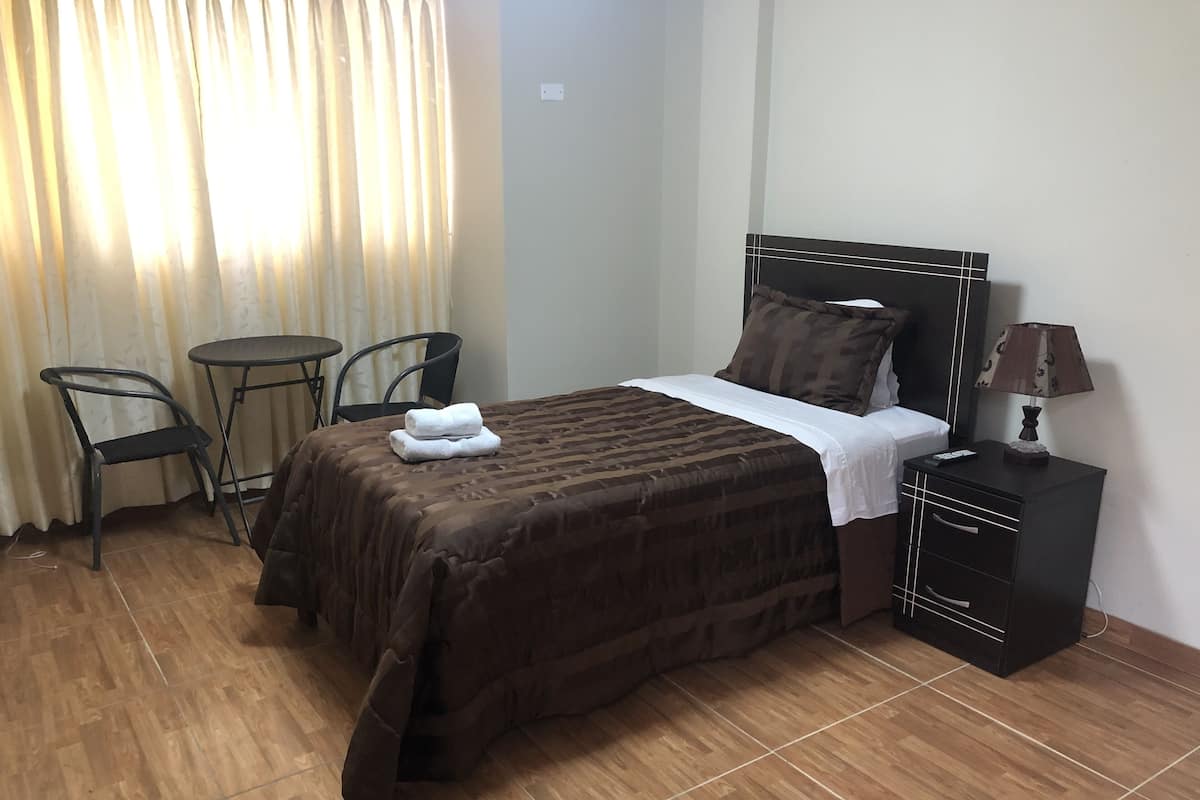 A neatly made bed with a brown cover, a bedside table with a lamp, a chair, and a small table.