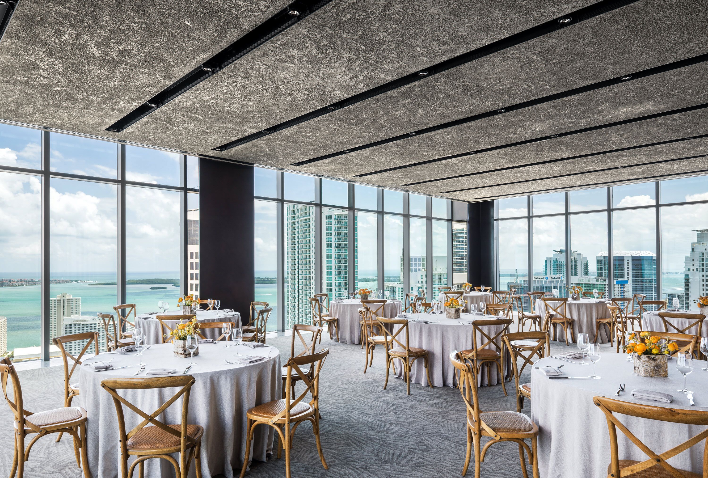 A spacious dining area with round tables and chairs, large windows offering a view of the city and ocean.