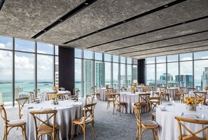 A spacious dining area with round tables and chairs, large windows offering a view of the city and ocean.