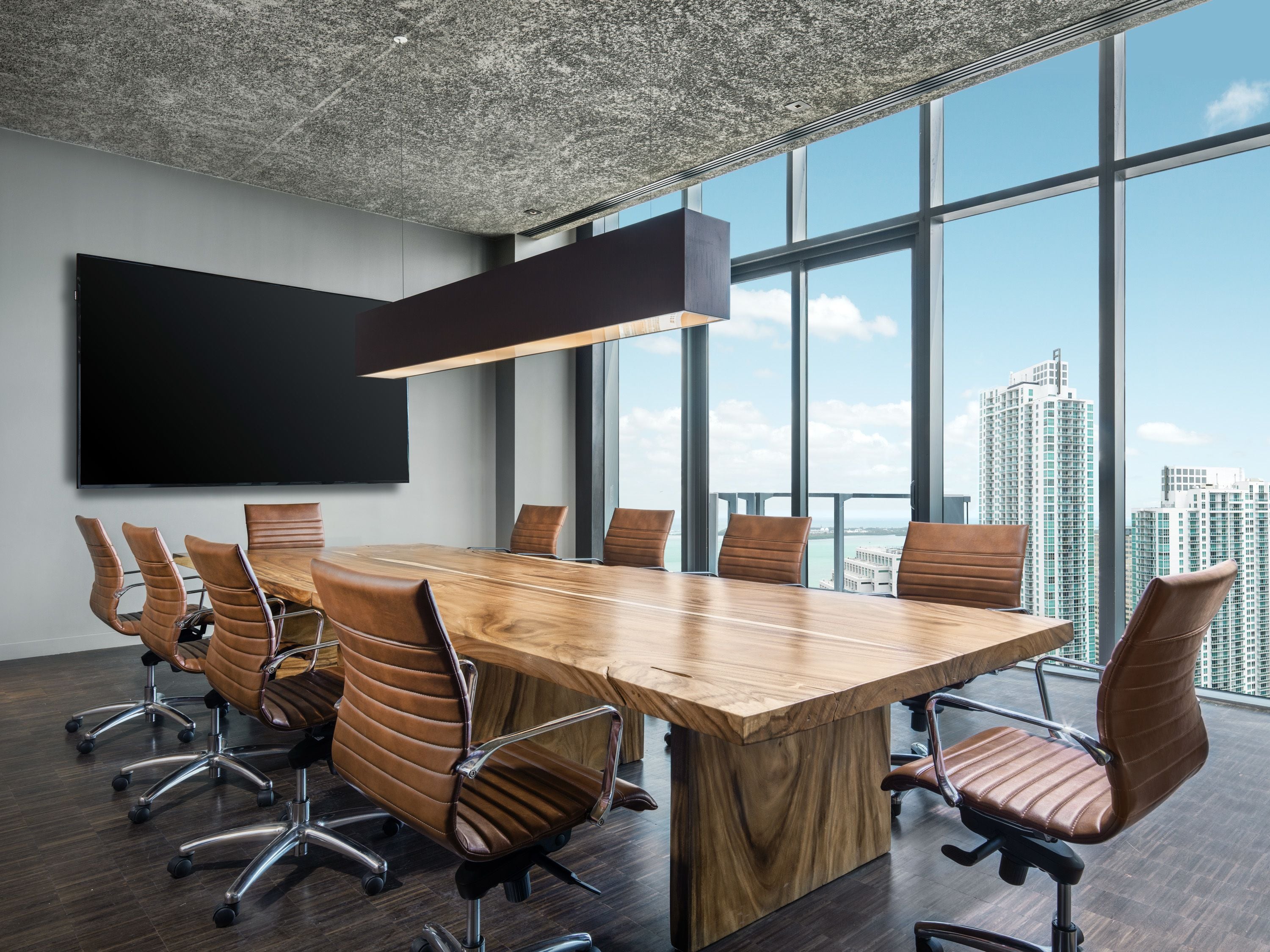 A conference room with a large wooden table, leather chairs, a mounted TV, and large windows offering a city view.