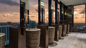 Un baño moderno con varios lavamanos y espejos grandes, con vistas a la ciudad a través de amplias ventanas.