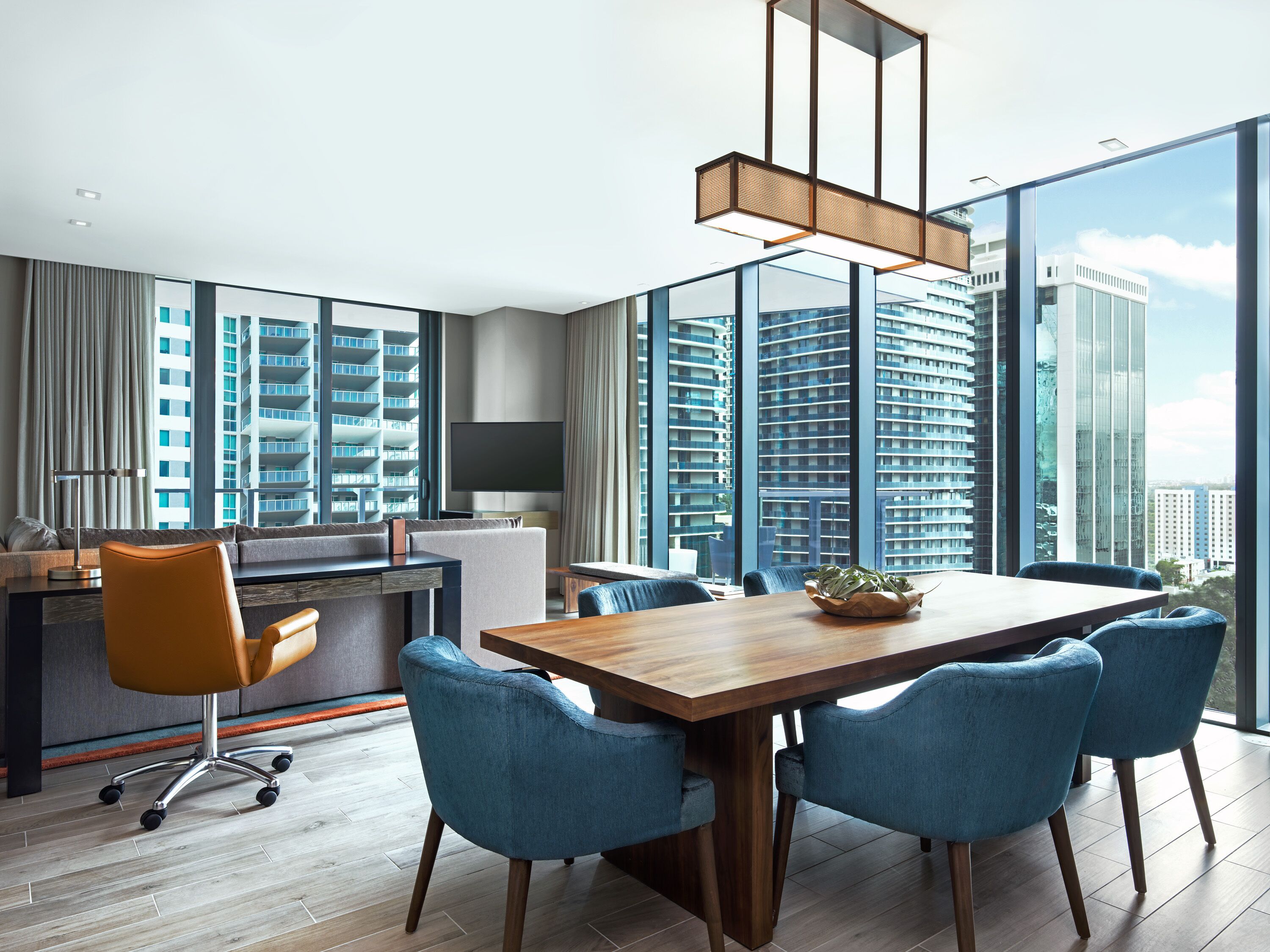 A modern hotel room with a large dining table, blue upholstered chairs, a wooden floor, a large window with a city view, and a flat-screen TV.