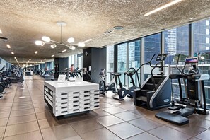 A modern gym with various cardio machines, a reception desk, and large windows offering views of the city.