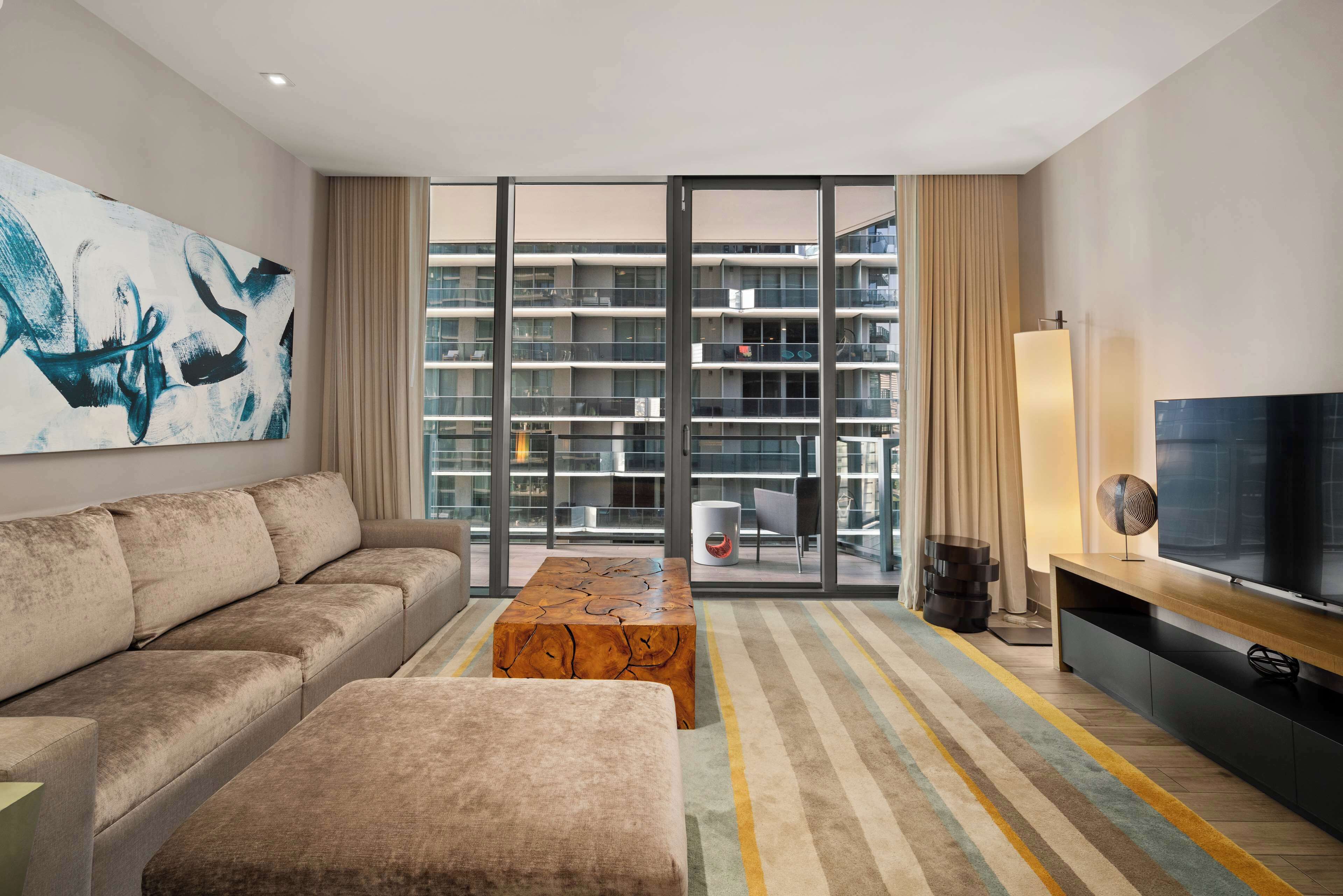 A modern living room with a sofa, ottoman, and a coffee table. There is a large flat-screen TV on a wooden stand, a floor lamp, and a painting on the wall. The room has large windows with a view of a building outside.