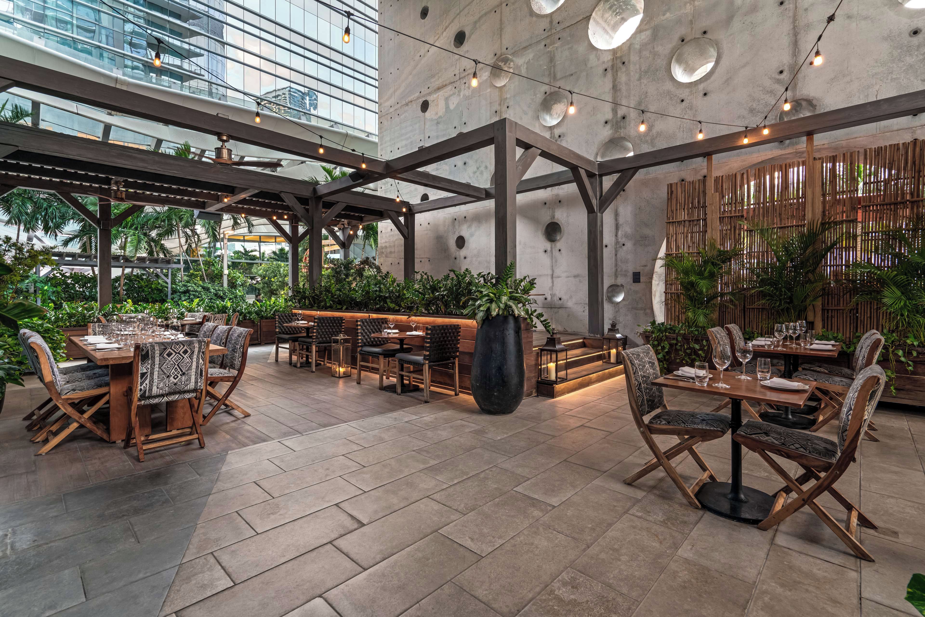 A spacious outdoor seating area with wooden tables and chairs, surrounded by potted plants and a concrete wall.