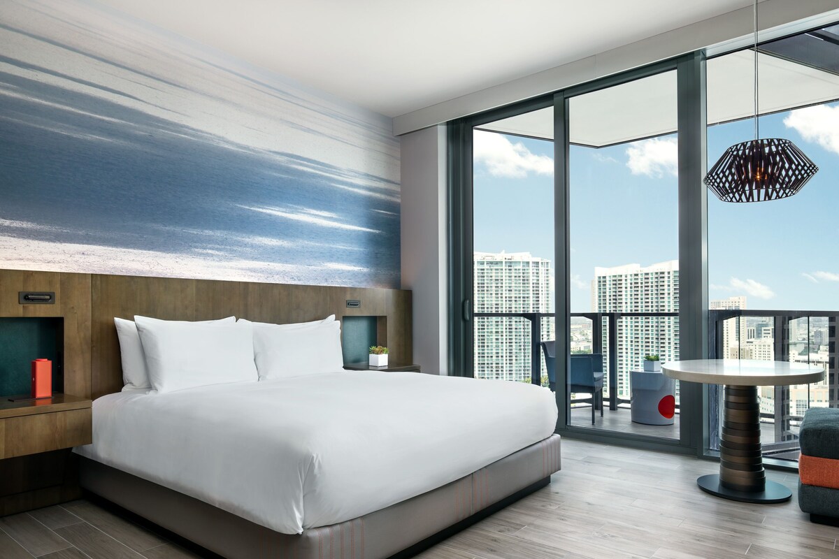 Una habitación de hotel moderna con una cama grande, vistas a la ciudad gracias a ventanas de gran altura y un balcón con mesa y sillas.