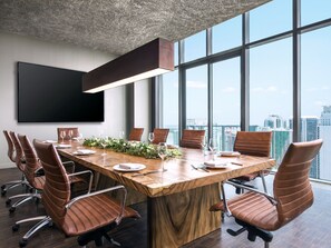 A conference room with a long table, brown leather chairs, a flat-screen TV, and a city view through large windows.