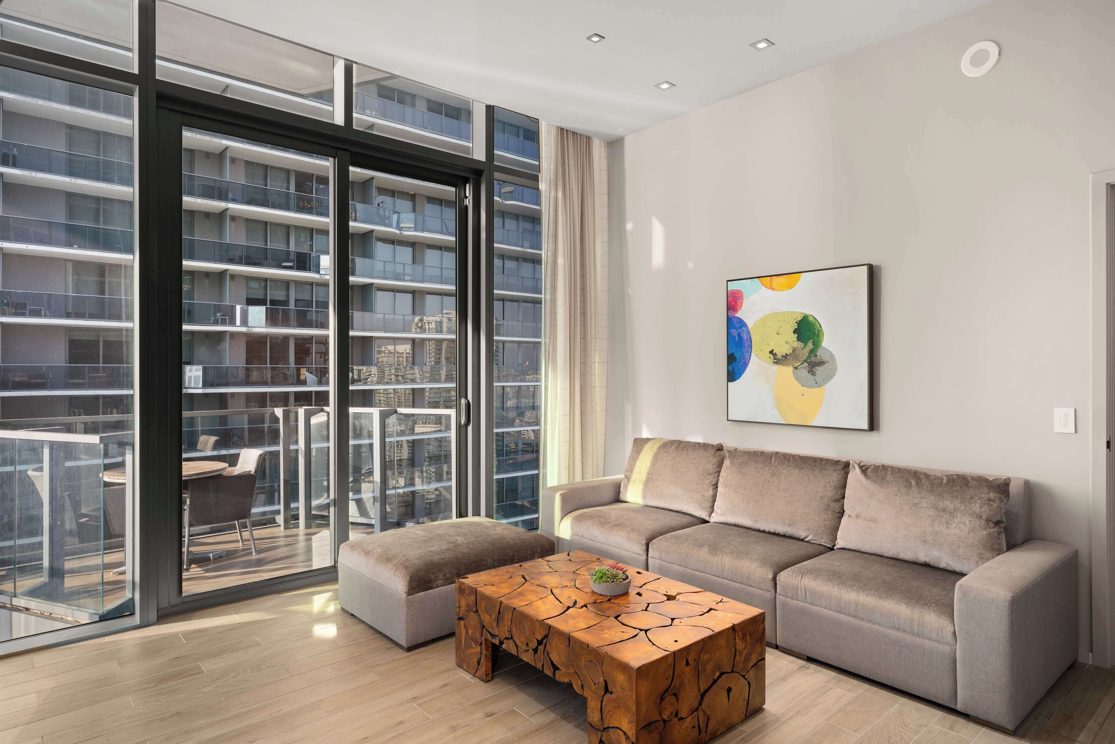 A modern living room with a grey sofa, a wooden coffee table, and a large window overlooking a cityscape.