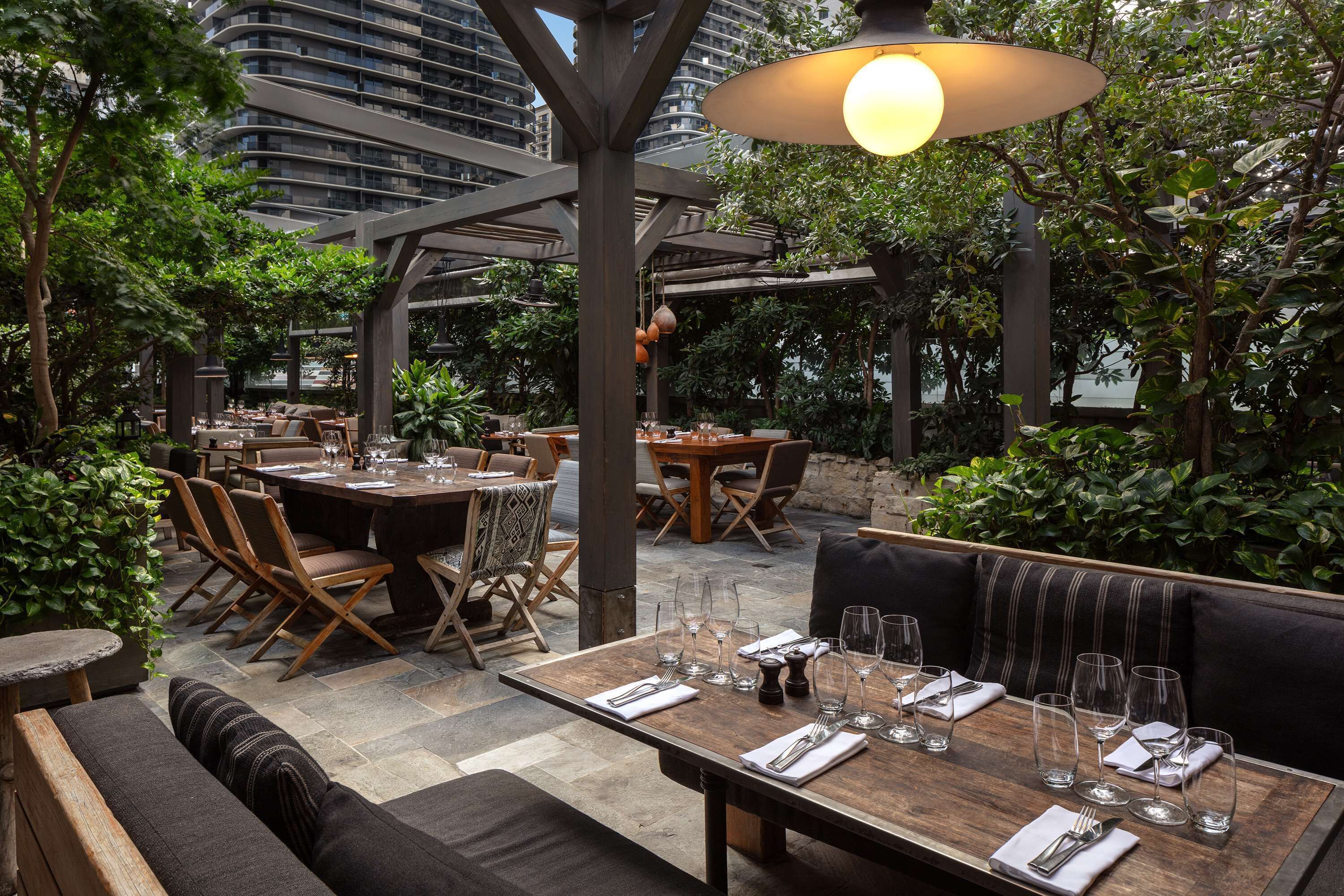 An outdoor dining area with multiple tables and chairs, surrounded by greenery and a modern building in the background.