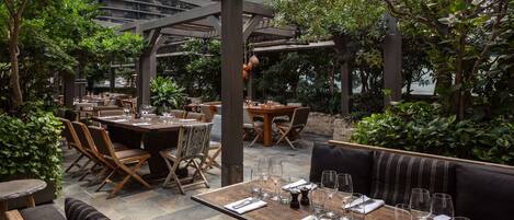 An outdoor dining area with multiple tables and chairs, surrounded by greenery and a modern building in the background.
