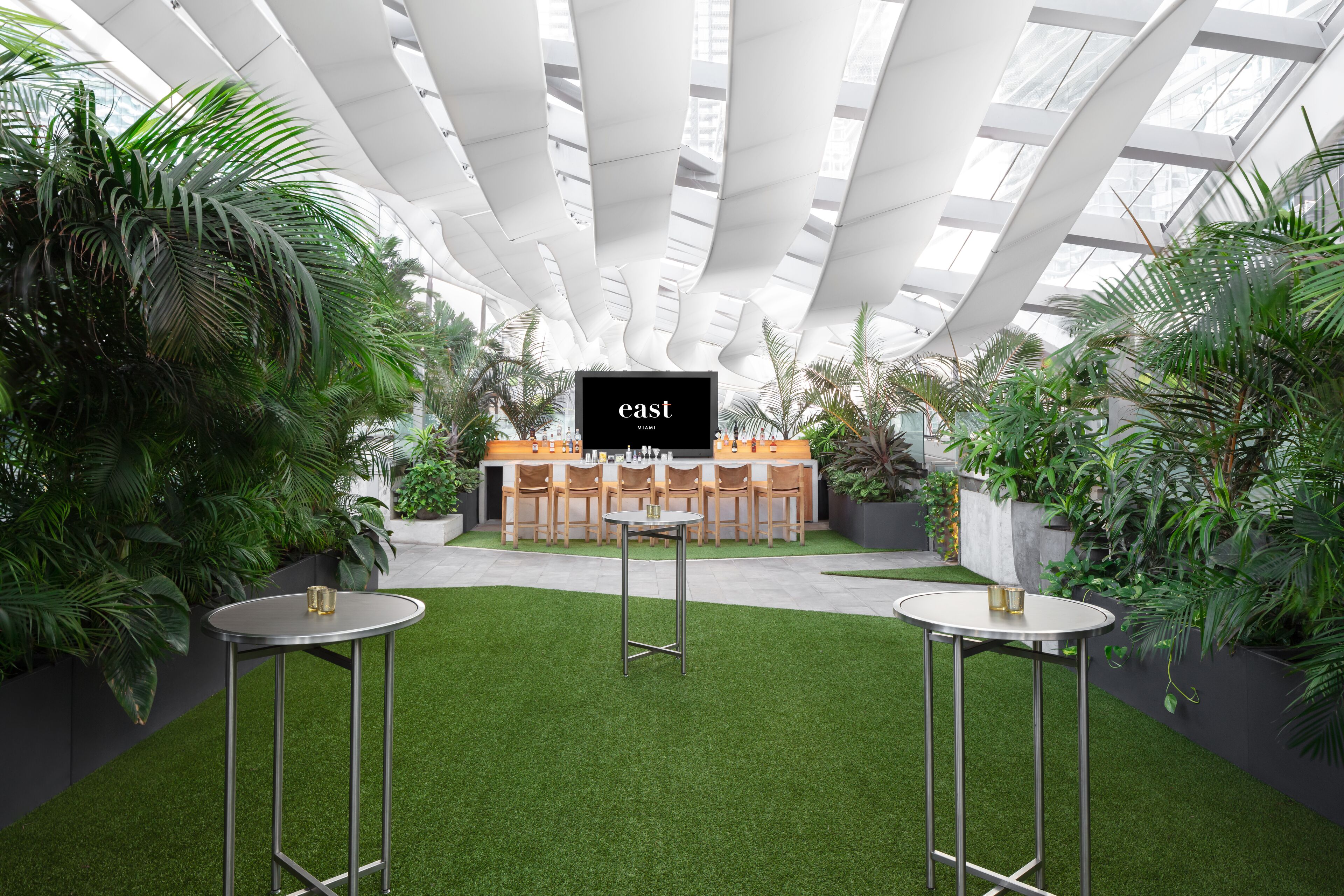 A modern indoor space with a central bar area, surrounded by lush greenery and a glass ceiling.