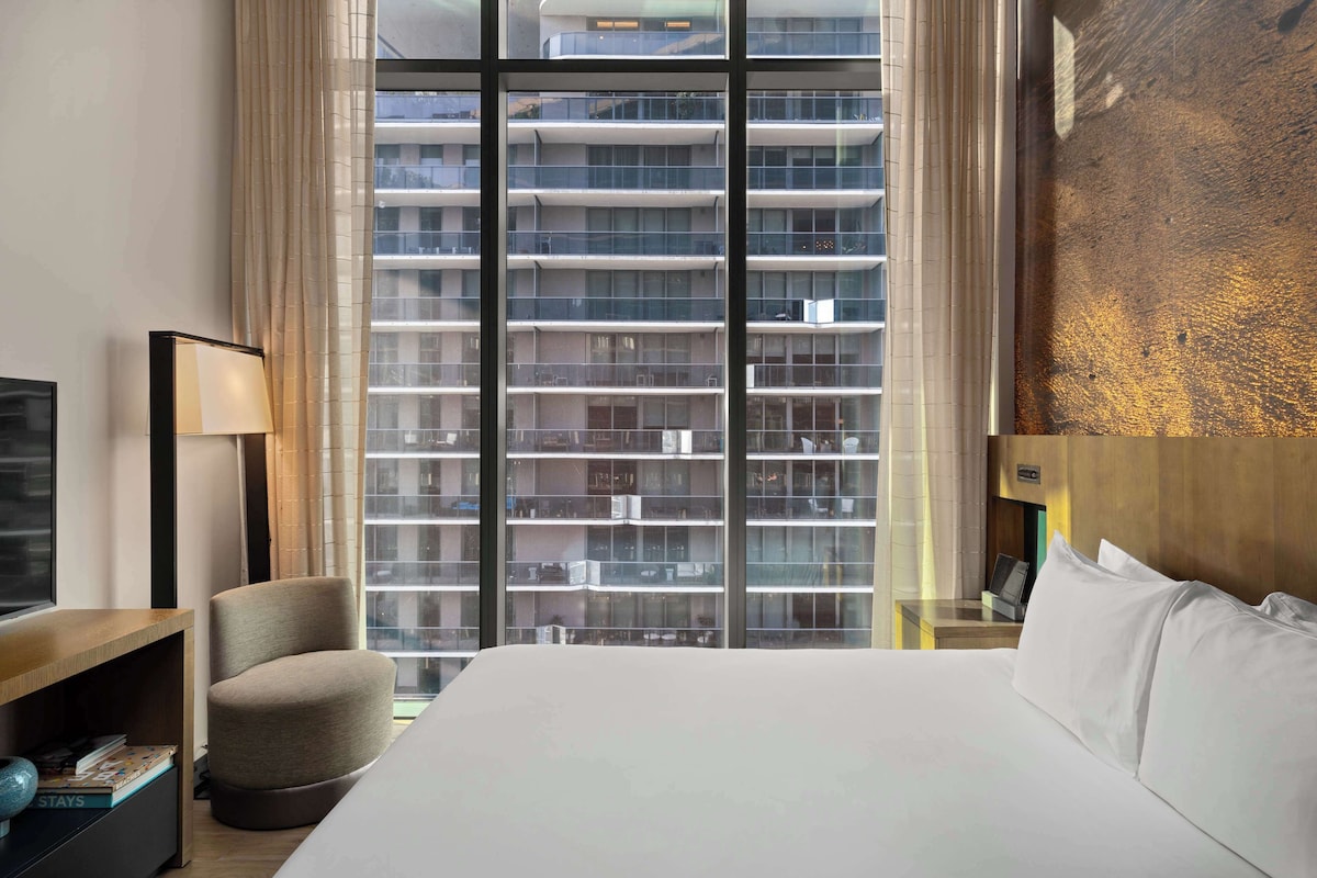 Habitación de hotel con una cama grande, una silla, un escritorio con lámpara y vistas a un edificio alto a través de ventanales.