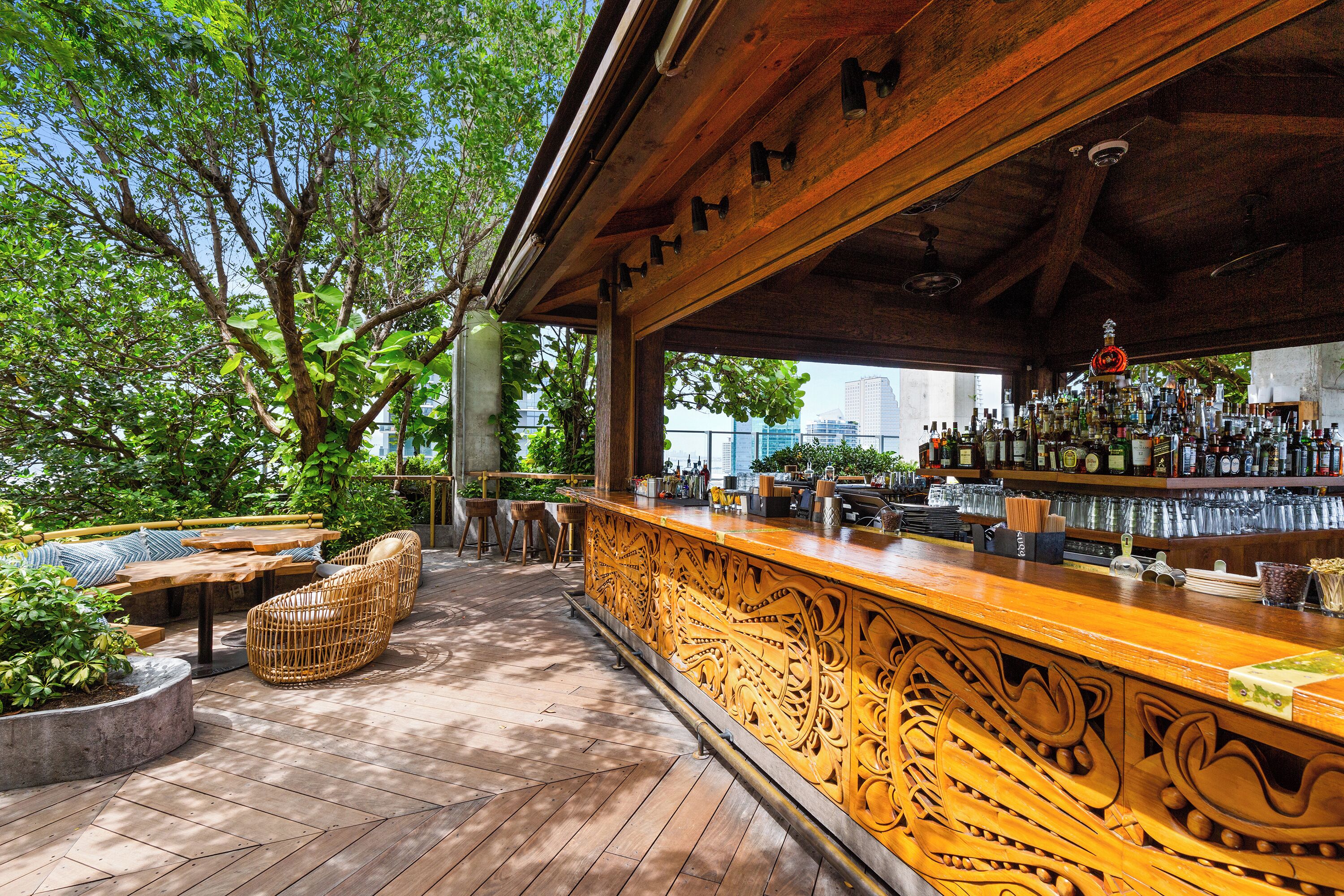 Zona de bar al aire libre con mobiliario de madera, una barra con intrincados grabados y vista al horizonte de la ciudad.