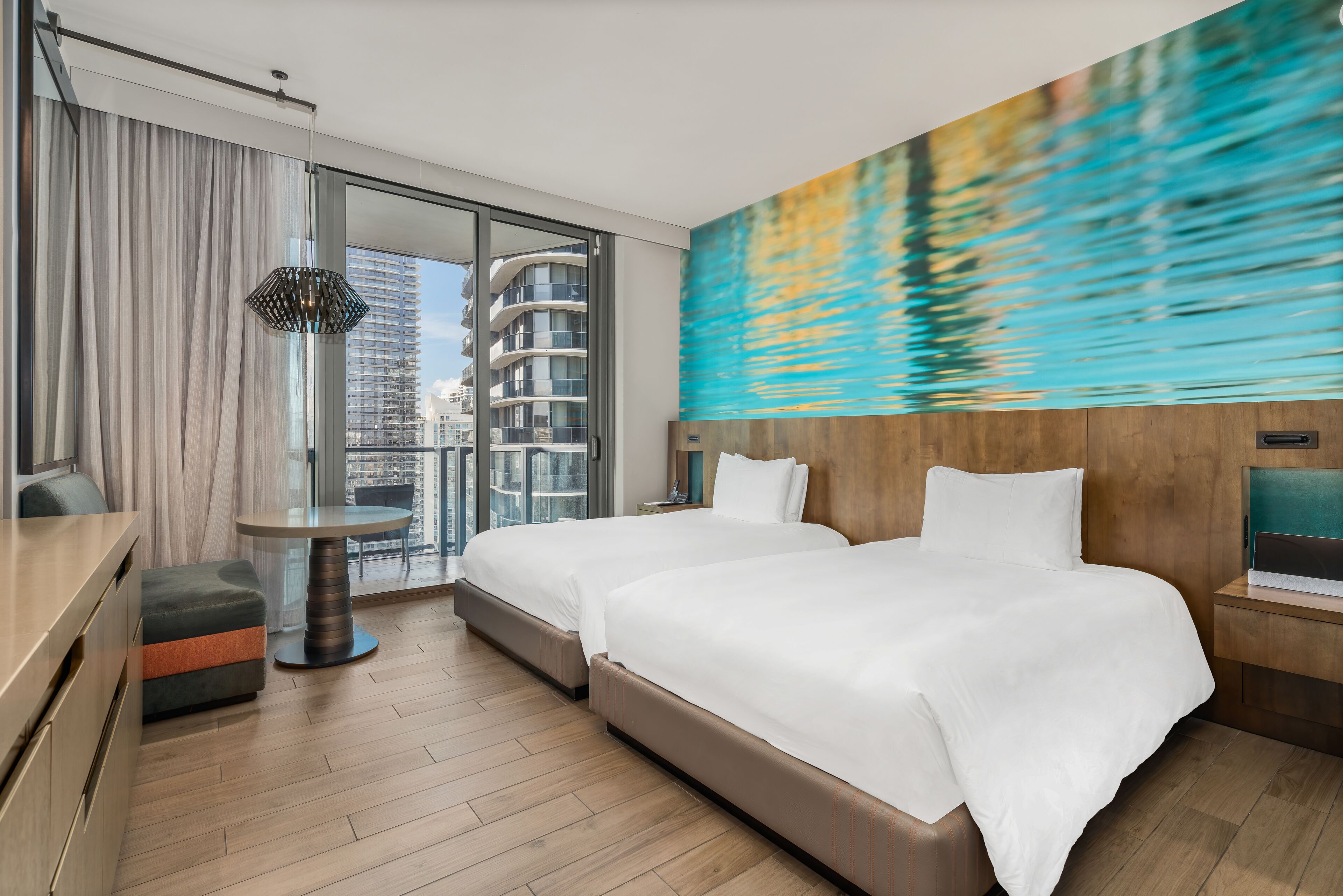 A modern hotel room with two beds, a dining table, and a large window with a city view.