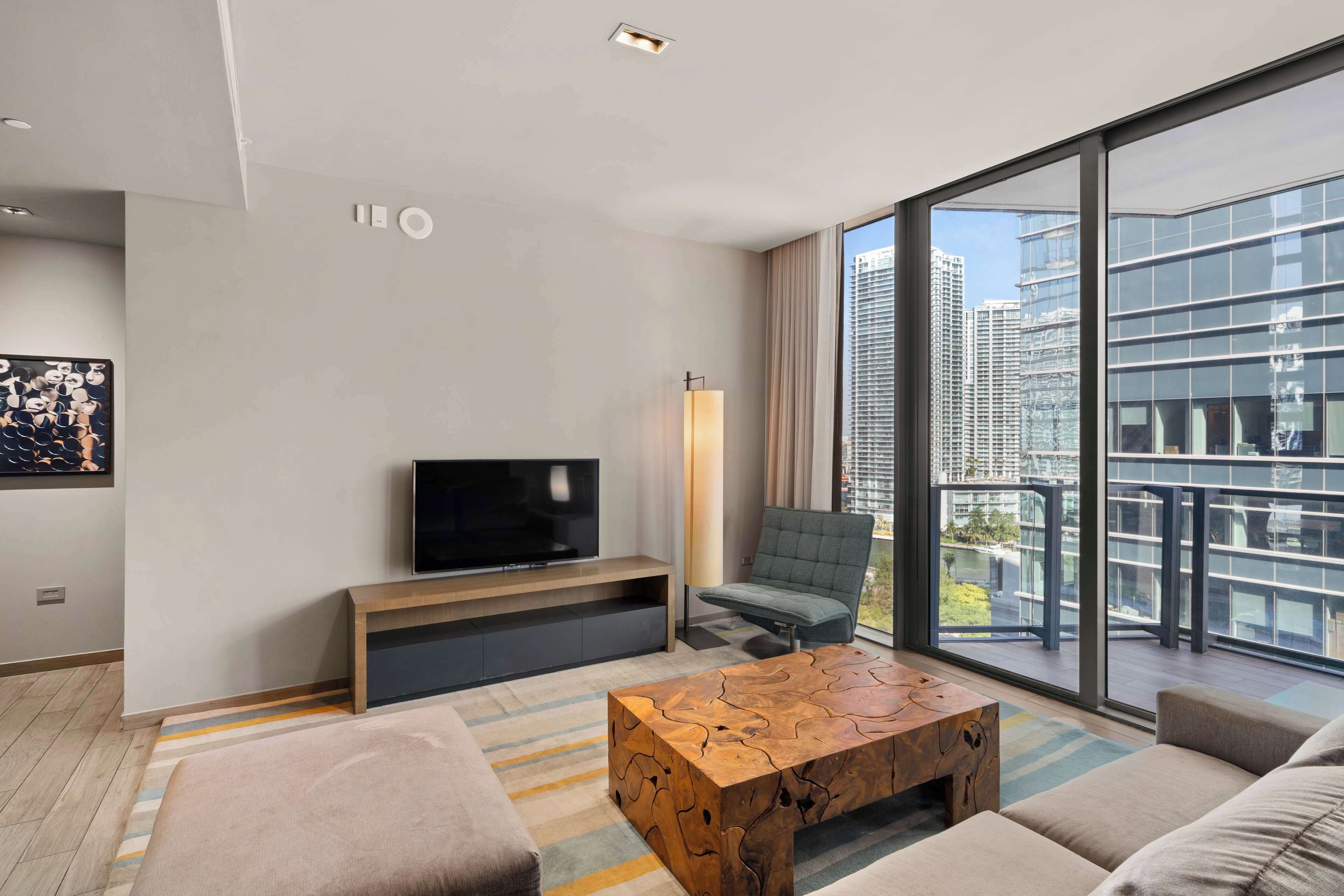 A modern living room with a flat-screen TV, a wooden coffee table, and a large window offering a city view.