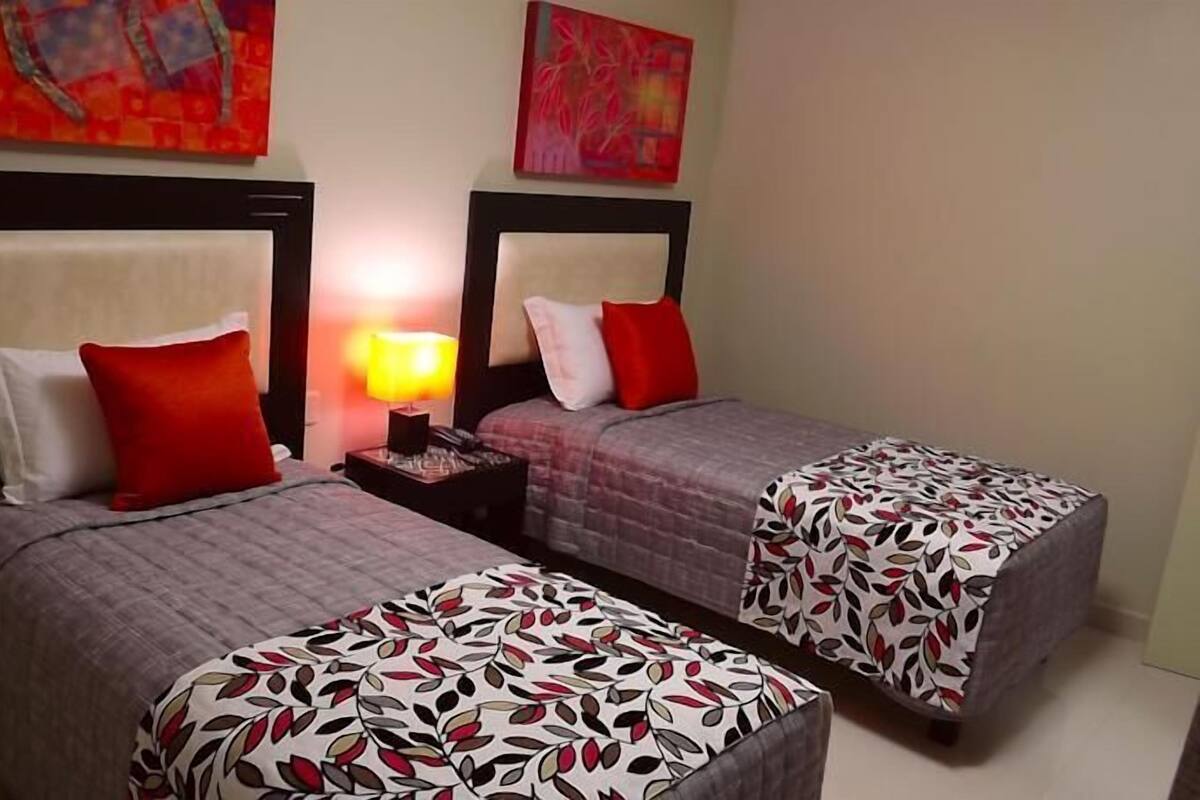 A hotel room with two single beds, each with a red pillow and a patterned bedspread. There is a nightstand with a lamp between the beds.