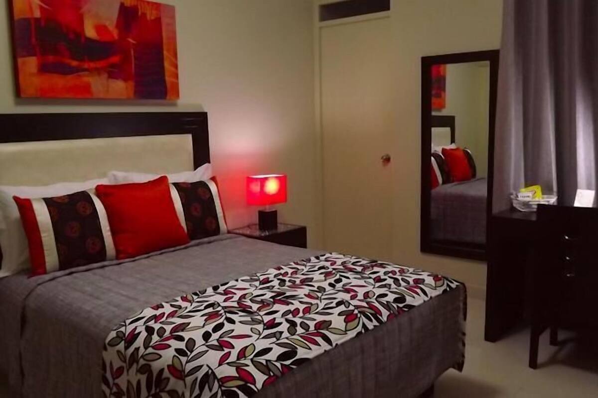 A hotel room with a bed, a bedside table with a lamp, a desk, a chair, a mirror, and a painting on the wall.
