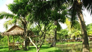 A lush green park with a thatched-roof pavilion and a calm pond. - Racha Herb Gardens (Saraphi)
