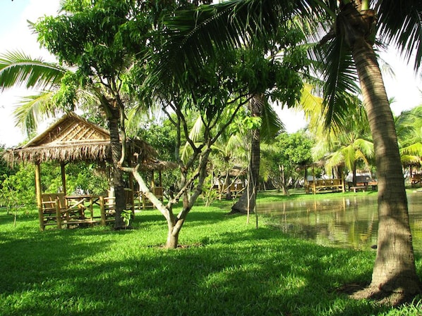 A lush green park with a thatched-roof pavilion and a calm pond.
