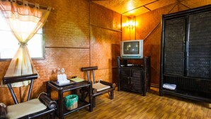 A room with a wooden floor, a television on a stand, a small table with a chair, and a window with a curtain. - Racha Herb Gardens (Saraphi)