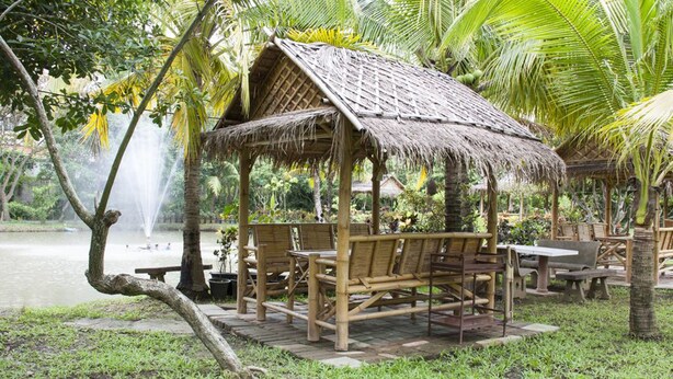 A thatched-roof gazebo with bamboo furniture, a pond, and palm trees. - Racha Herb Gardens (Saraphi)