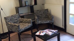 Un salon avec deux fauteuils à motifs, une table basse avec des magazines, une bibliothèque et un radiateur.
