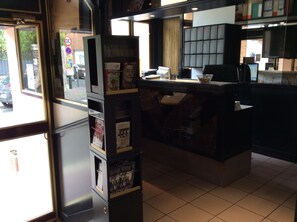 A reception area with a counter, a display of magazines, and a view of the street outside. - Hotel De La Place (Malakoff)