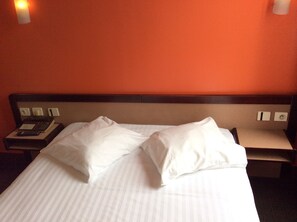 A hotel room with a bed, pillows, a nightstand with a phone, and a wall-mounted light. - Hotel De La Place (Malakoff)