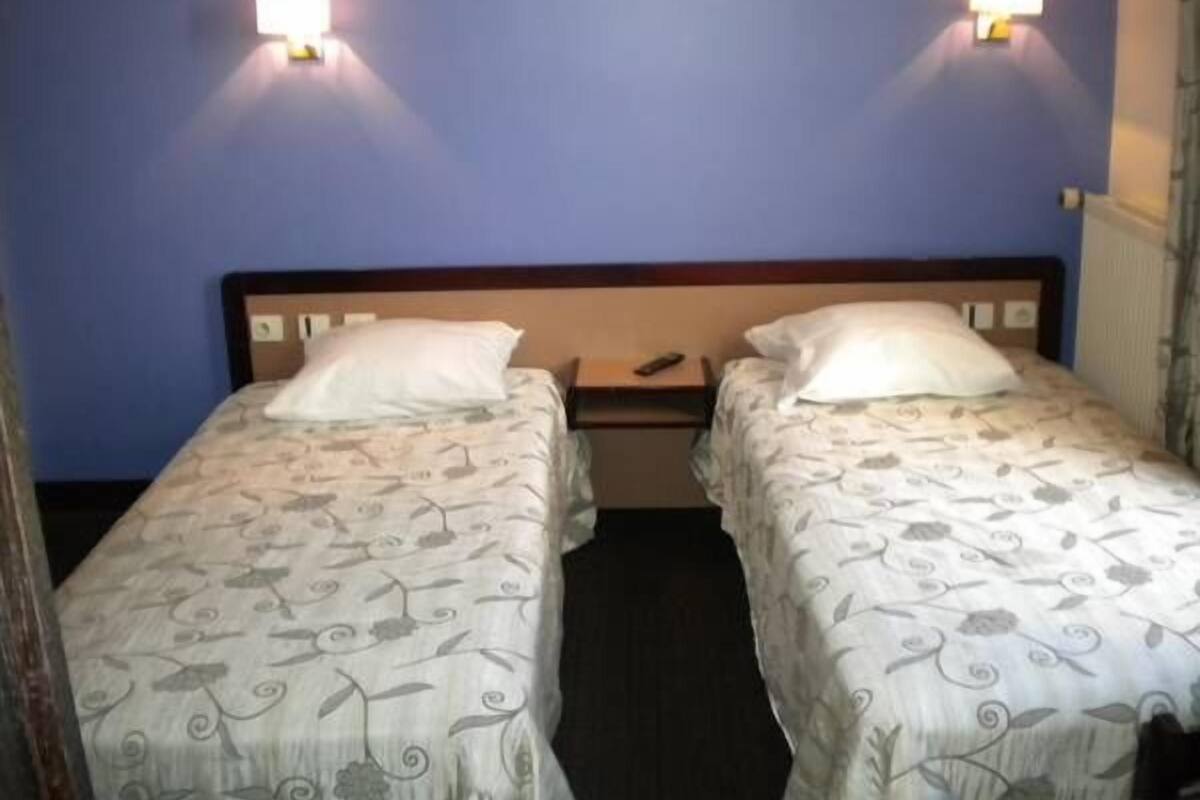 A twin room with two single beds, each with a white floral bedspread, a wooden headboard, and a bedside table. There are two wall-mounted night lights on either side of the bed.