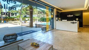 Un moderno vestíbulo de hotel con una recepción, una zona de piscina visible a través de puertas de cristal y un entorno exterior tropical.