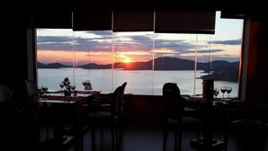 Ein Essbereich mit großem Fenster und Blick auf einen Sonnenuntergang über dem Wasser, mit Tischen und Stühlen für das Essen eingerichtet.