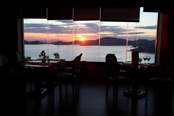 Un espace repas doté d’une grande fenêtre donnant sur un magnifique coucher de soleil sur une étendue d’eau, avec des chaises et des tables disposées pour les repas.