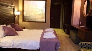 Ein Hotelzimmer mit Bett, Nachttisch, Lampe, Fenster mit Jalousien, Kommode und Spiegel.