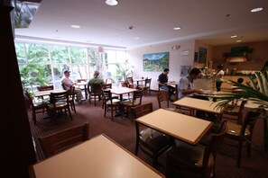 A restaurant with multiple tables and chairs, large windows with a view of greenery, and a kitchen area visible in the background. - Metrotel Express (San Pedro Sula)
