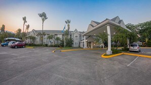 A hotel with a covered entrance, parking lot, and palm trees. - Metrotel Express (San Pedro Sula)
