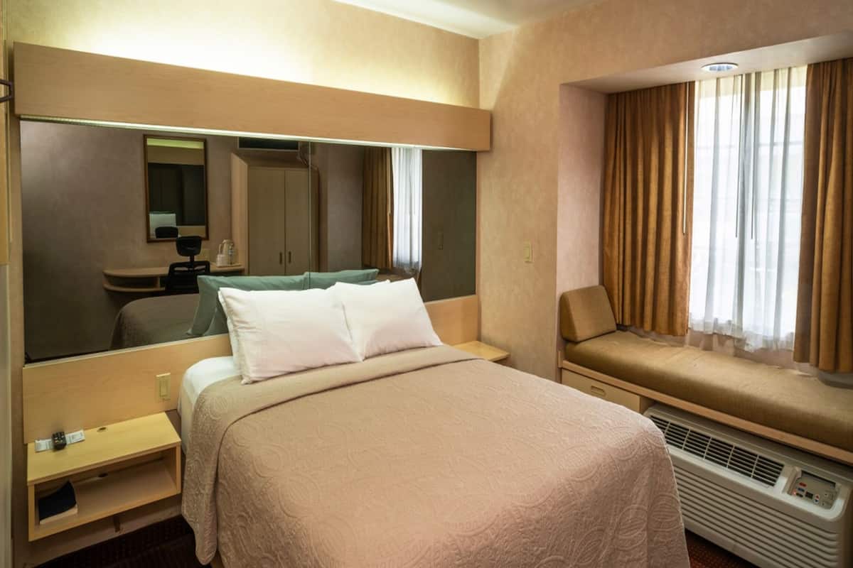 Habitación de hotel con cama, escritorio, silla, espejo y ventana con cortinas.
