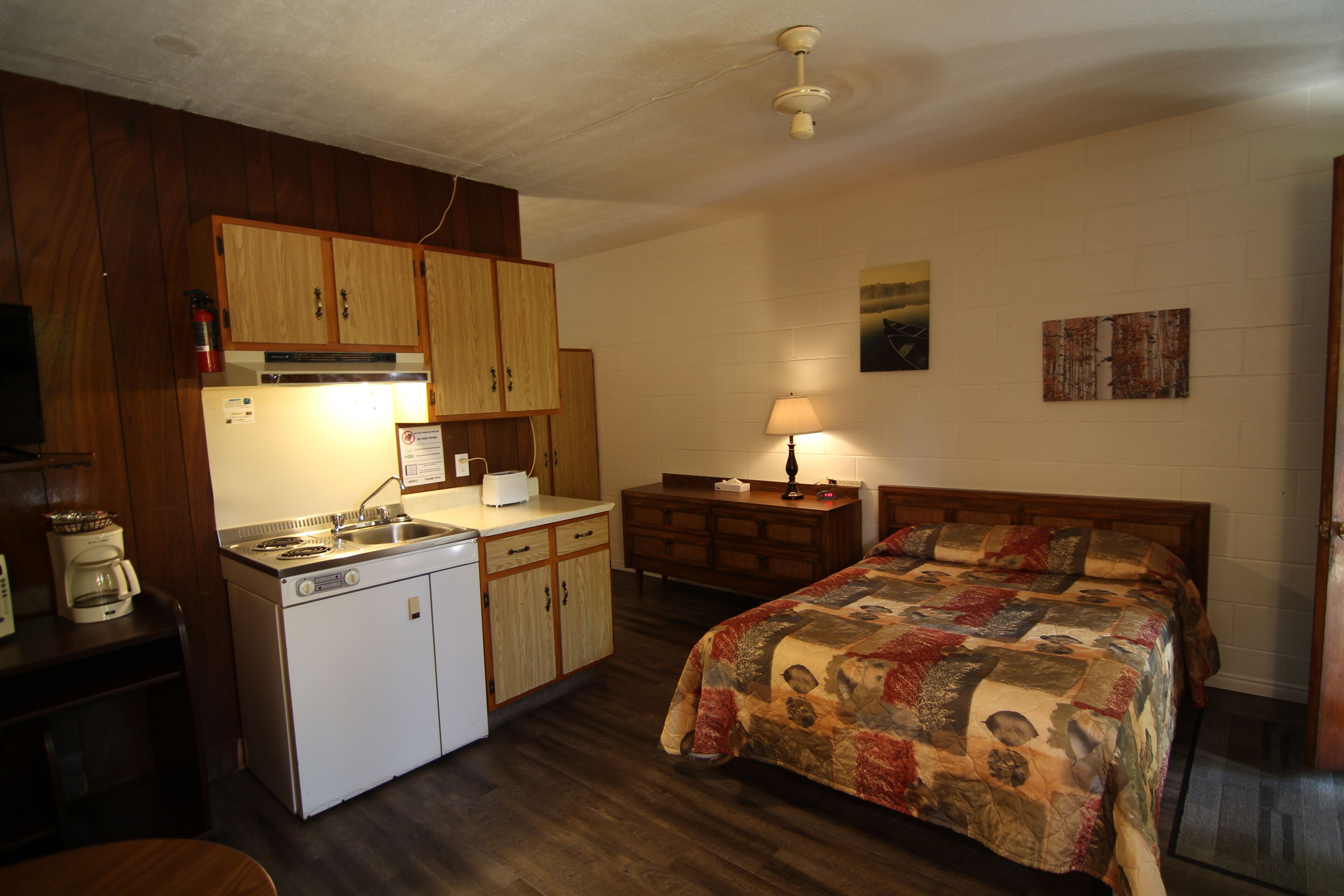Habitación compacta con armarios de madera, una cama con colcha estampada, una pequeña kitchenette con refrigerador, estufa y lavaplatos, y un ventilador de techo.