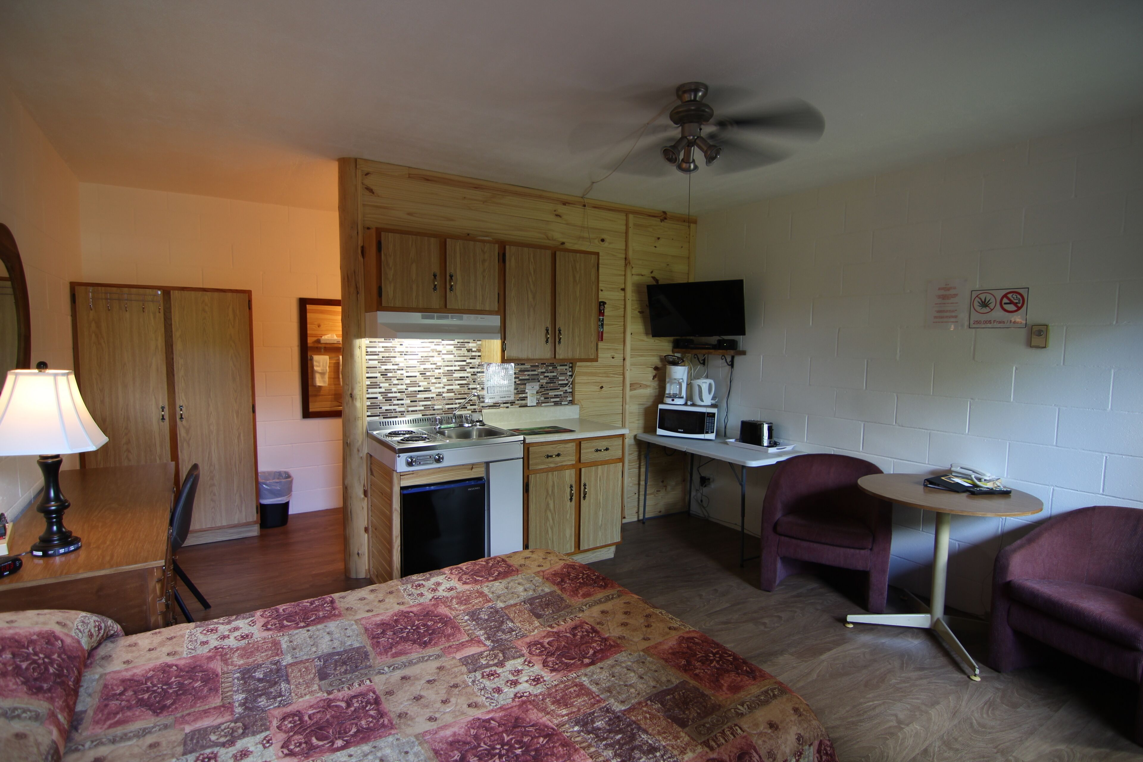 Une chambre d’hôtel compacte avec une kitchenette, un lit recouvert d’une courtepointe à motifs, deux fauteuils violets, une petite table et un ventilateur de plafond.