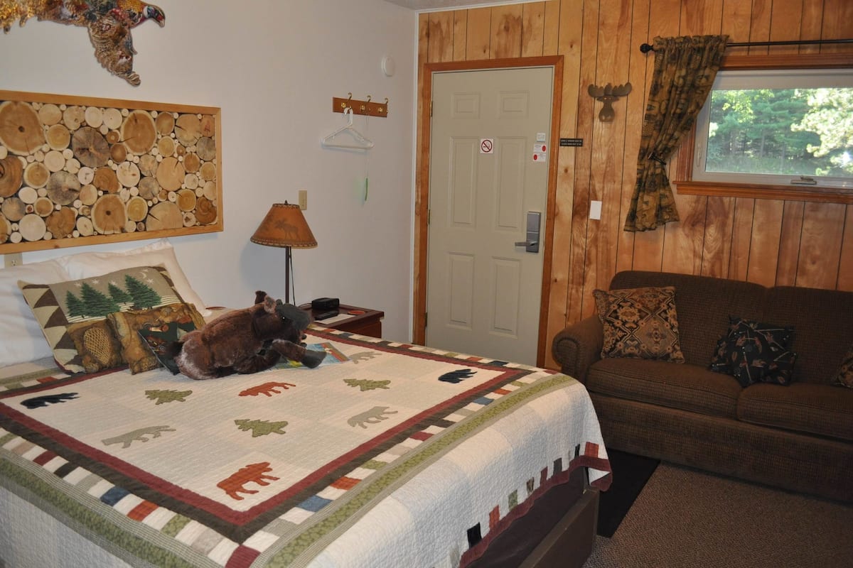 A cozy room with a bed, a couch, a quilt with animal patterns, a wooden wall, a door, and a window with a view of trees.