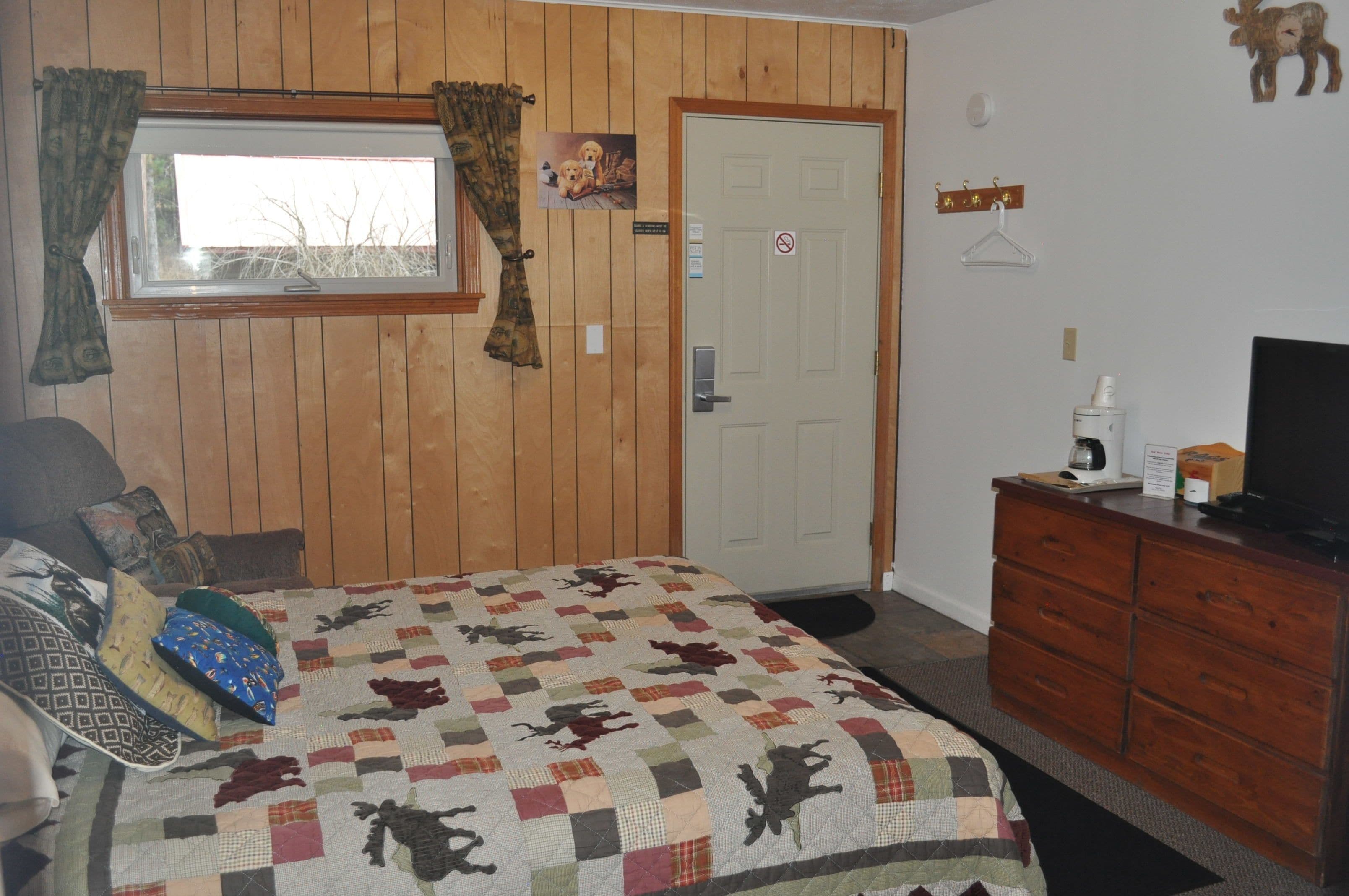 Une chambre avec un mur lambrissé en bois, un lit recouvert d’une courtepointe, une commode, un téléviseur et une porte avec une pancarte.