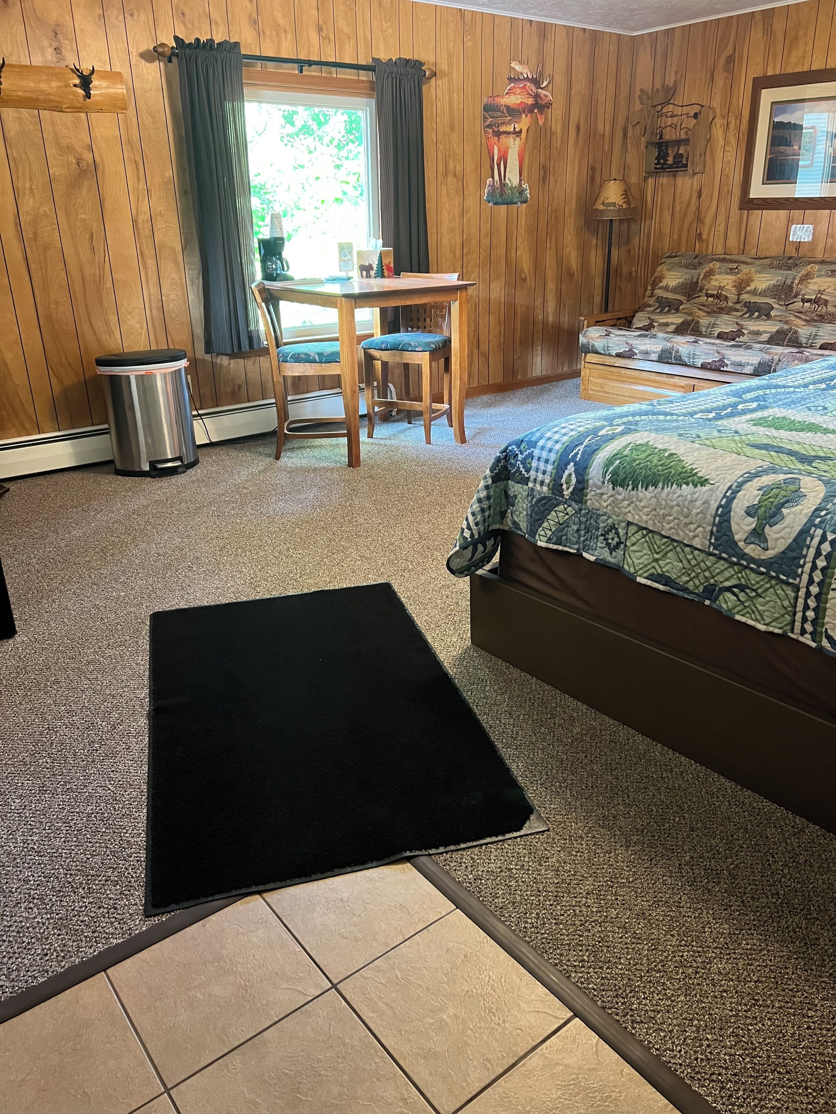 A room with a bed, a desk, a chair, a trash can, and a window with curtains.