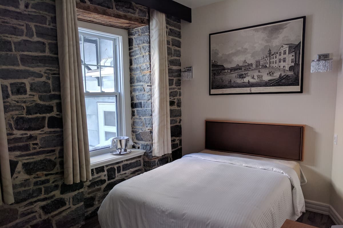 A bedroom with a stone wall, a window with curtains, a bed with a wooden headboard, and a framed picture on the wall.