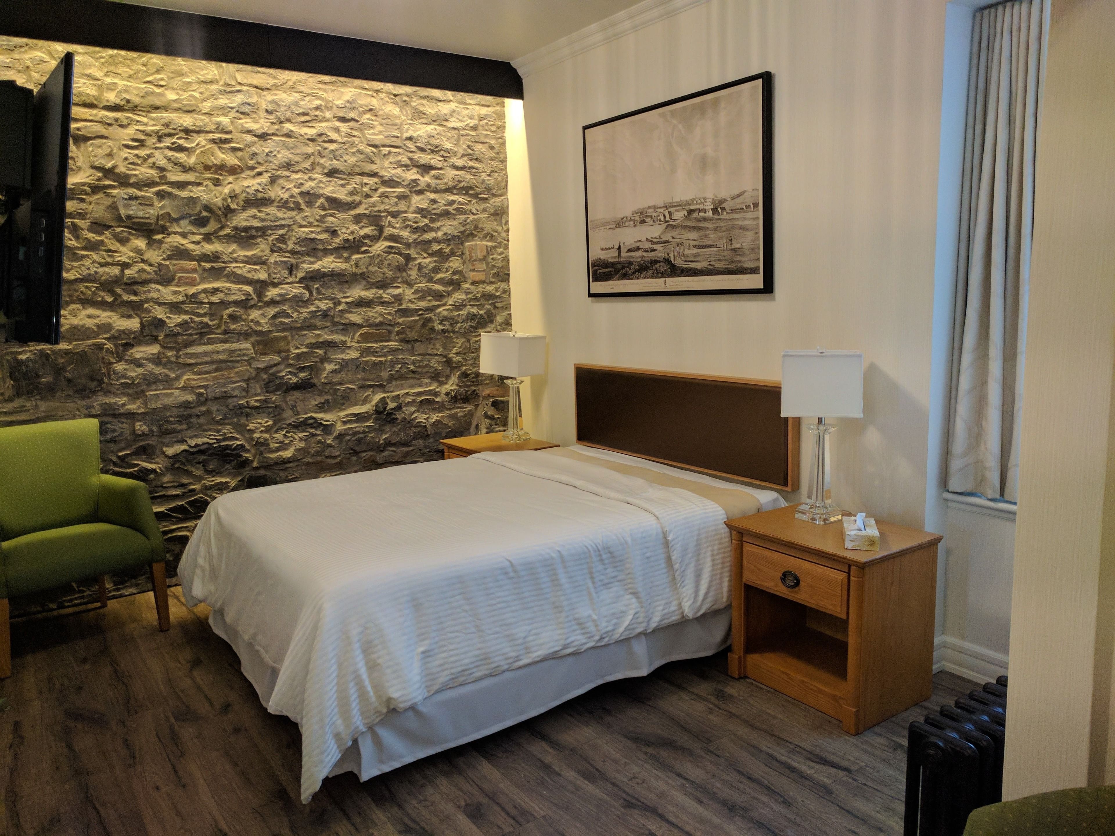 A hotel room with a stone wall, a bed with white bedding, a green chair, a wooden nightstand with a lamp, and a framed picture on the wall.