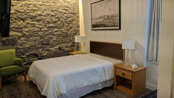 Quarto de hotel com parede de pedra, cama com roupa de cama branca, cadeira verde, mesa de cabeceira de madeira com abajur e quadro na parede.