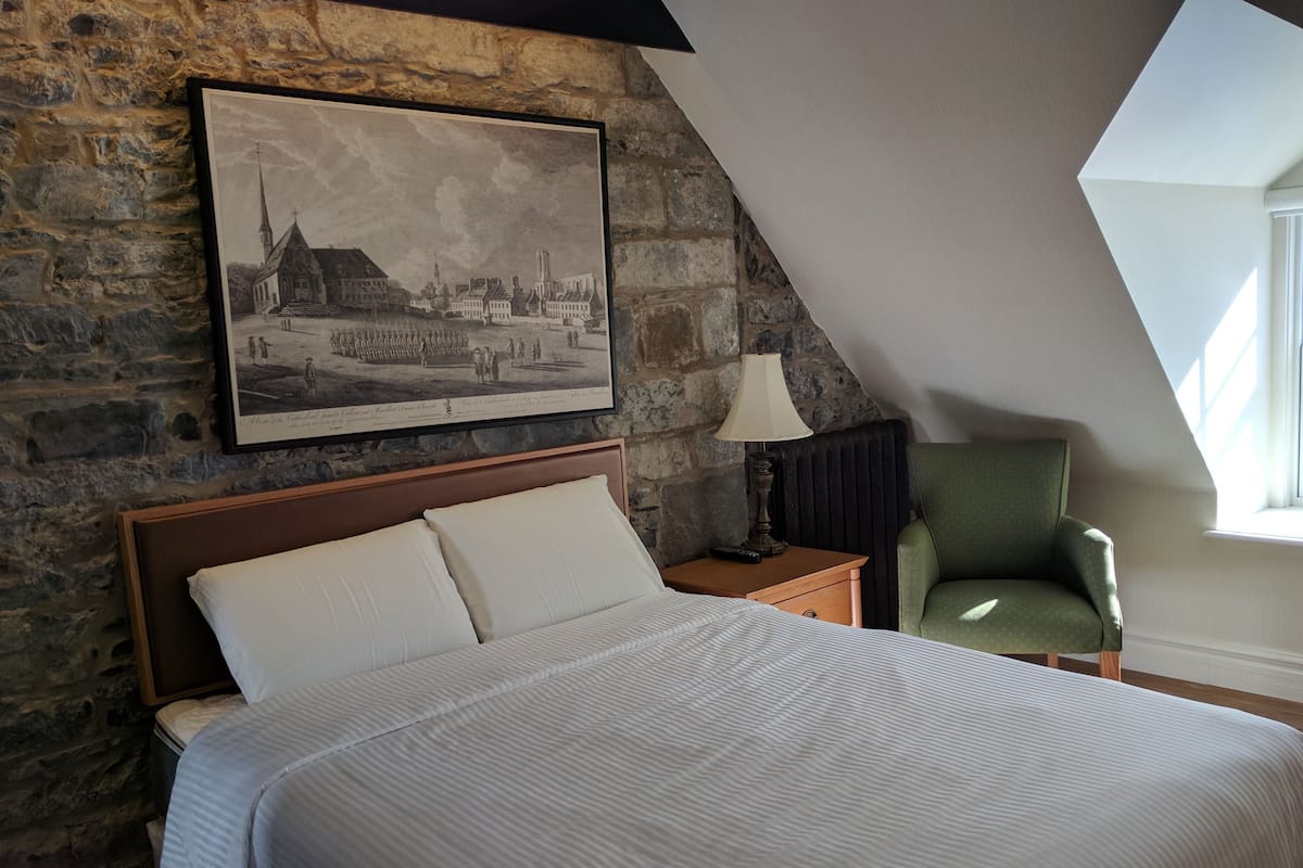 A bedroom with a stone wall, a bed with white linens, a framed historical photograph, a nightstand with a lamp, and a green armchair.