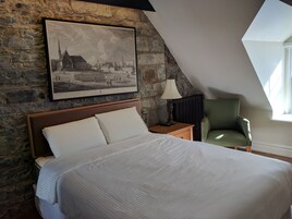 Un dormitorio con pared de piedra, una cama con ropa blanca, una fotografía histórica enmarcada, una mesita de noche con lámpara y un sillón verde.