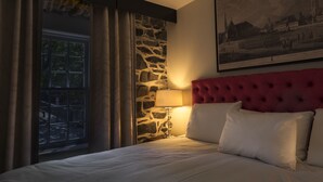 A hotel room with a stone accent wall, a large window with curtains, a red upholstered headboard, and a framed picture on the wall. - Auberge du Tresor (Québec City)