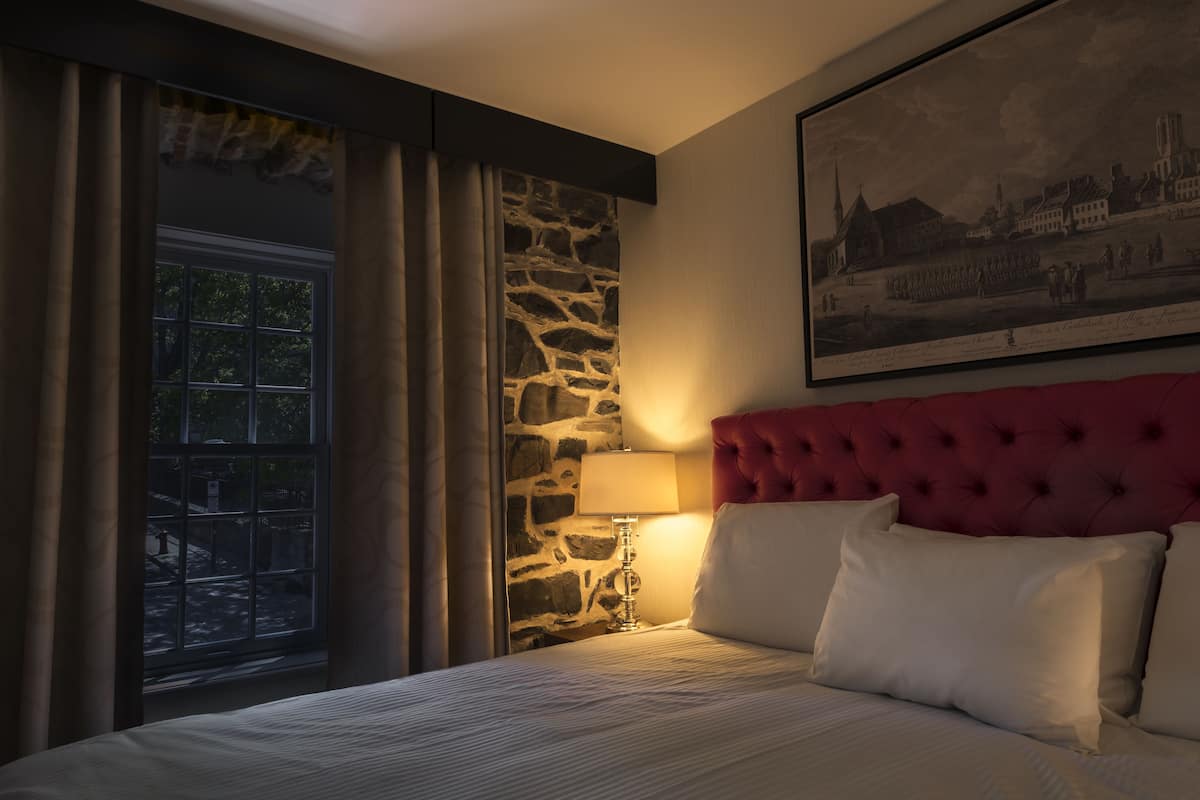 A hotel room with a stone accent wall, a large window with curtains, a red upholstered headboard, and a framed picture on the wall.