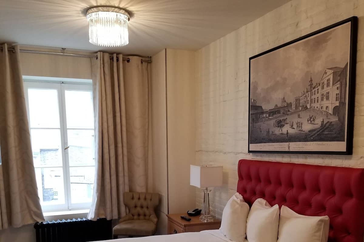 A large bed with white linens, a red upholstered headboard, a wooden nightstand with a lamp, a chair, and a framed historical painting on the wall.