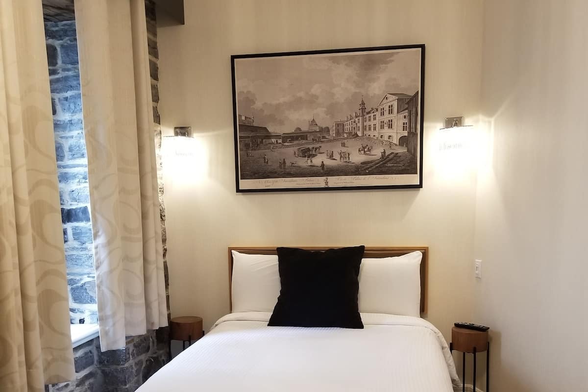 A hotel room with a large bed, a stone wall, a painting of a town scene, and a nightstand with a lamp.